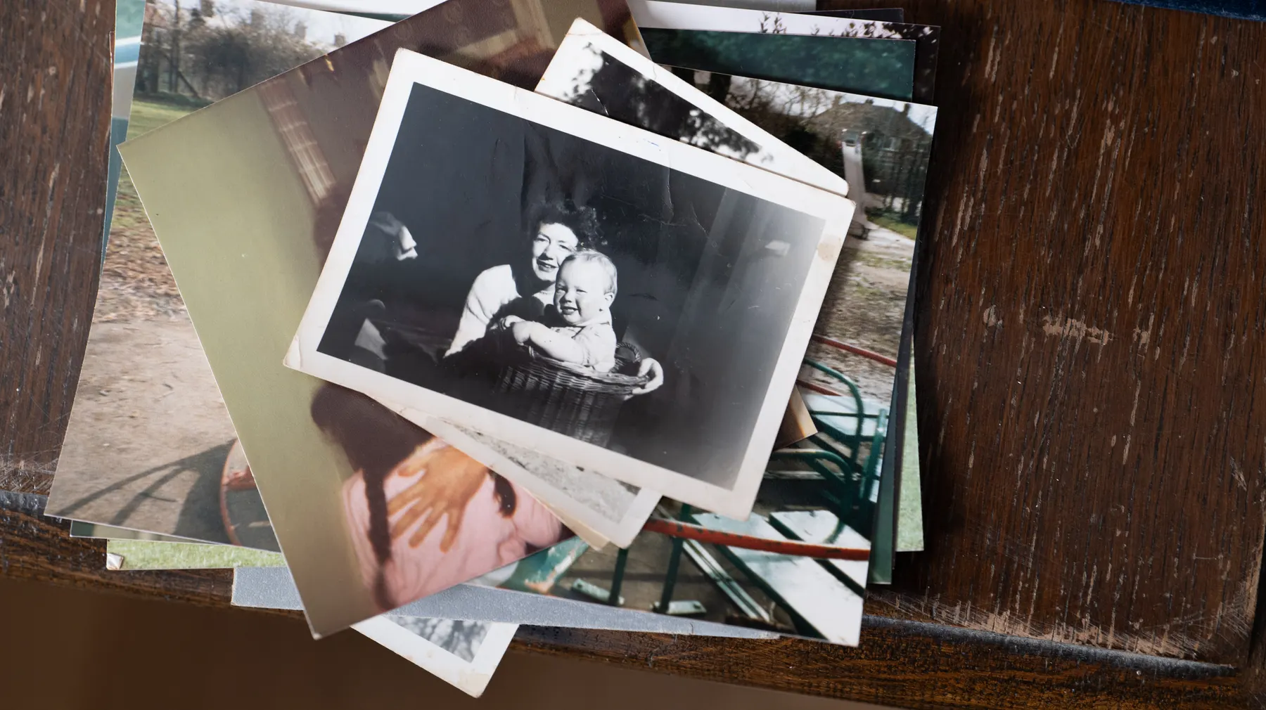 A stack of old family photographs on a wooden table, the top one of a baby in a wicker basket