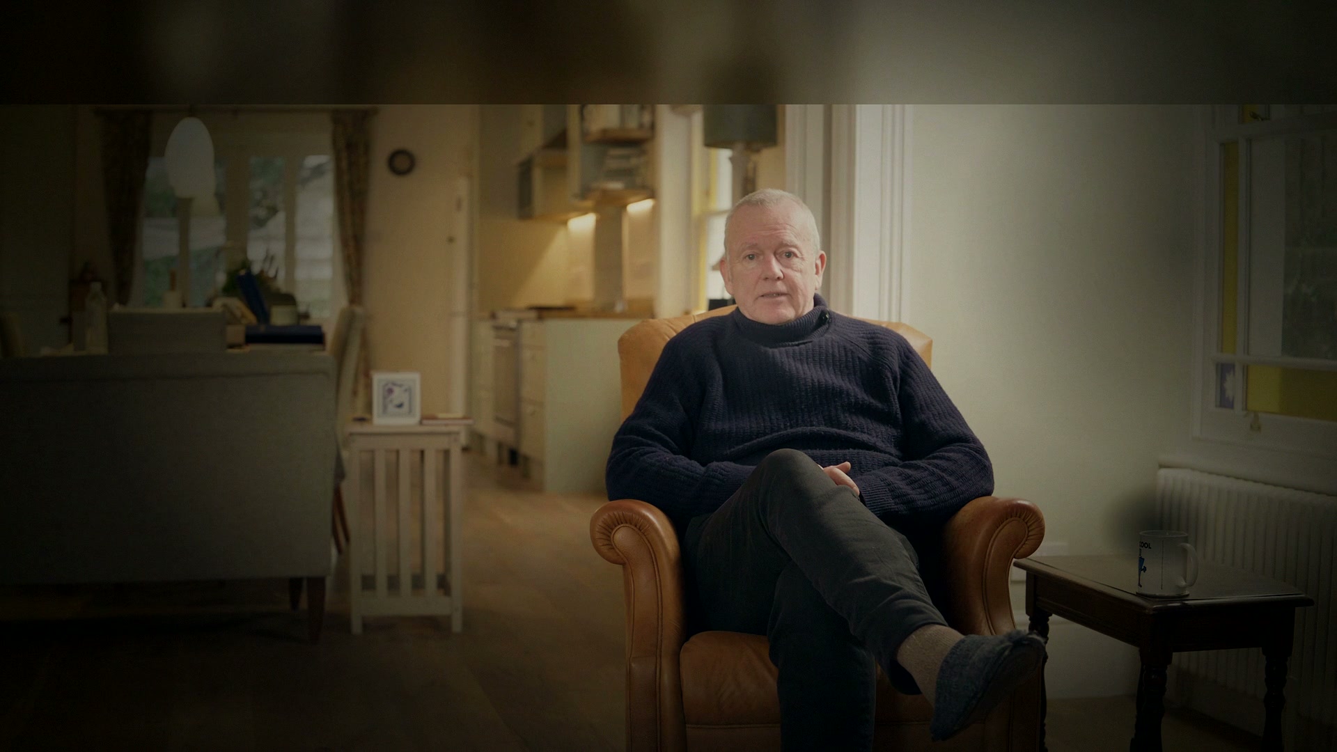 A man in a navy jumper sits in a leather armchair in his living room, listening