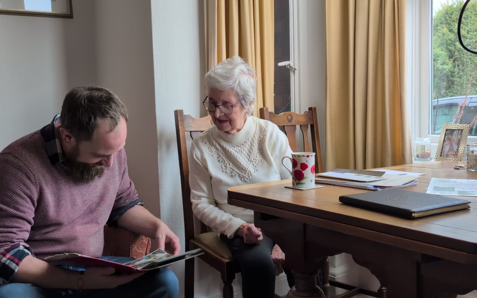 A grandmother at her dining table looks through old photographs with her grandson before filming